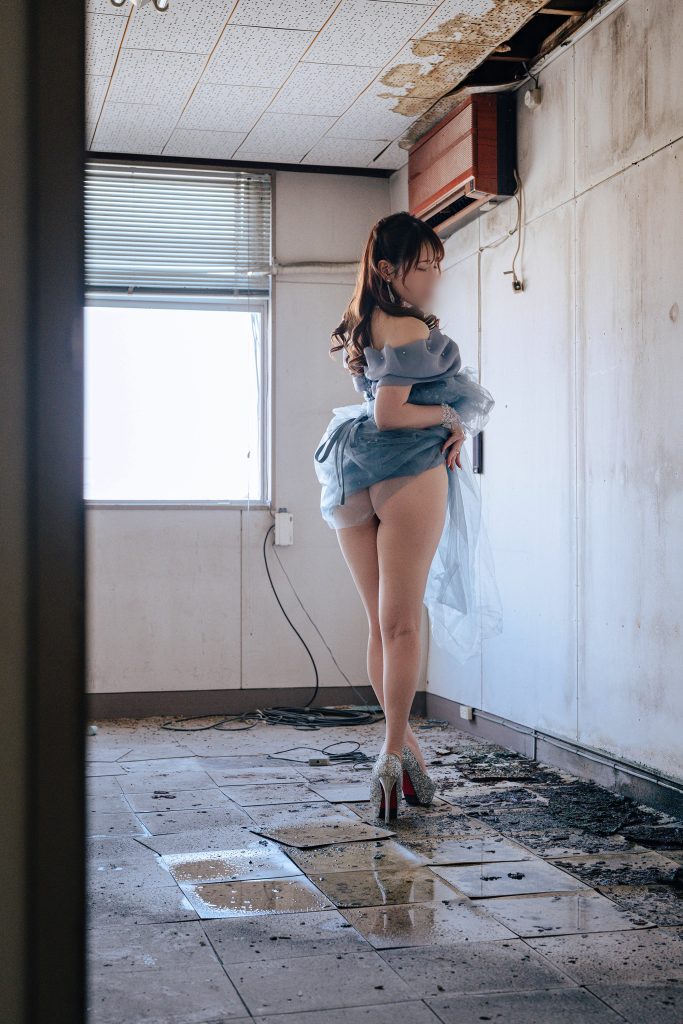 フォトグラファー赤木友厚による、福岡の廃墟スタジオ『朝日陸運大川営業所』での撮影。青いドレスを着たモデルAOIさんが、朽ちた部屋の大きな窓を背にして立ち、振り返るポーズでドレスの裾を持っている。窓からの強いバックライトが彼女の輪郭を強調し、ガレキの床や朽ちた壁が退廃美を演出。ちゃんみな『ハレンチ』 MV再現ポートレート。顔にはぼかし処理。