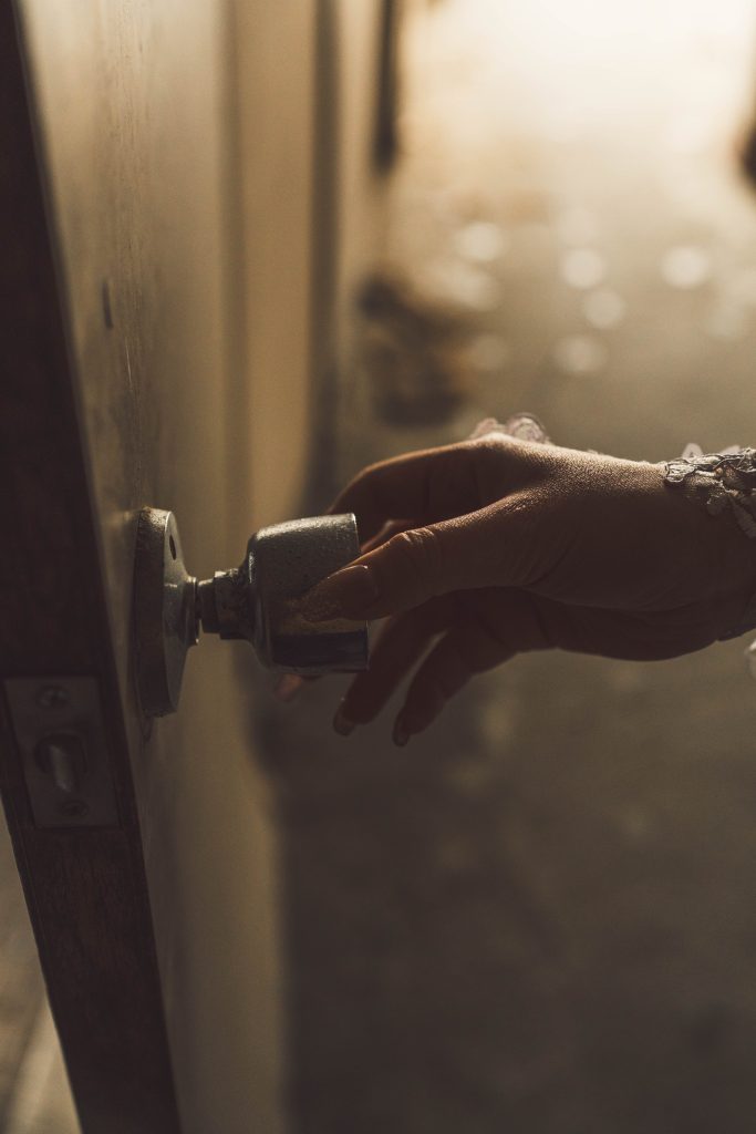 フォトグラファー赤木友厚が、福岡の廃墟スタジオ『朝日陸運大川営業所』で撮影したポートレートのクローズアップ。レースのグローブを着用したモデルAOIさんの手が、古いドアのノブを掴んで開けようとしている。ドアの向こう側からは強い光が差し込み、ガレキが見える。ちゃんみな『ハレンチ』MV再現。茶色っぽいトーン。