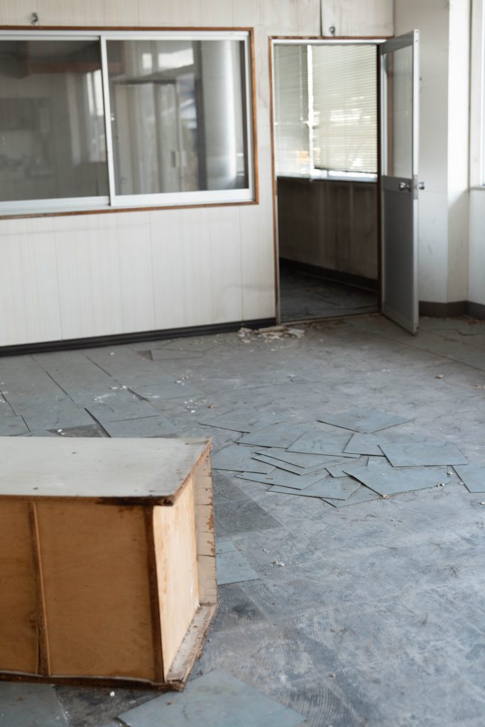 朝日陸運大川営業所の明るい廃墟室内。剥がれたタイルが床に散らばり、手前には古い木のカウンターがある。開かれたドアから差し込む自然光が美しい、清潔感のある廃墟ロケーション。フォトグラファー赤木友厚によるロケハン。