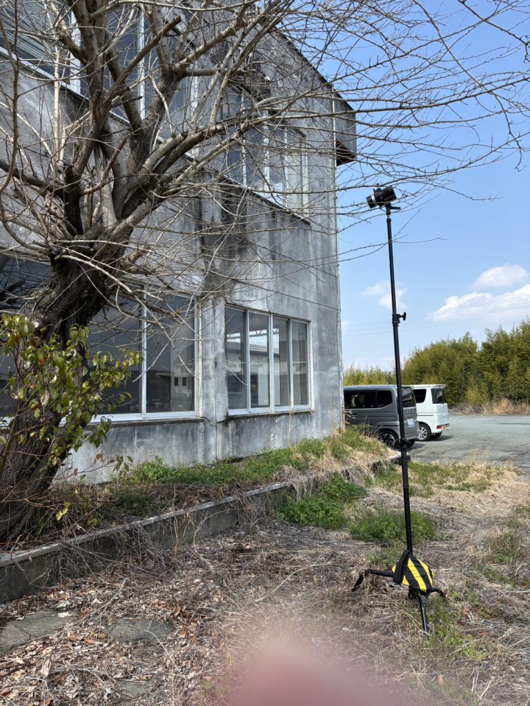 フォトグラファー赤木友厚が、福岡の廃墟スタジオ『朝日陸運大川営業所』でロケハンを行っている様子。コンクリートの廃墟ビルの前に、カメラが設置された黒い高い三脚ポールが立ち、ウェイト用の黄色と黒の砂袋が置かれている。枯れ木と草が茂り、奥に車が見える。