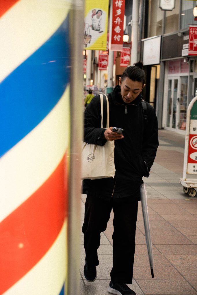 福岡市博多区の川端商店街にて、理髪店の赤・青・黄色のサインポール越しに、スマートフォンを見ながら歩く男性を捉えたストリートスナップ。背景には「博多弁番付」の赤い垂れ幕が見える。