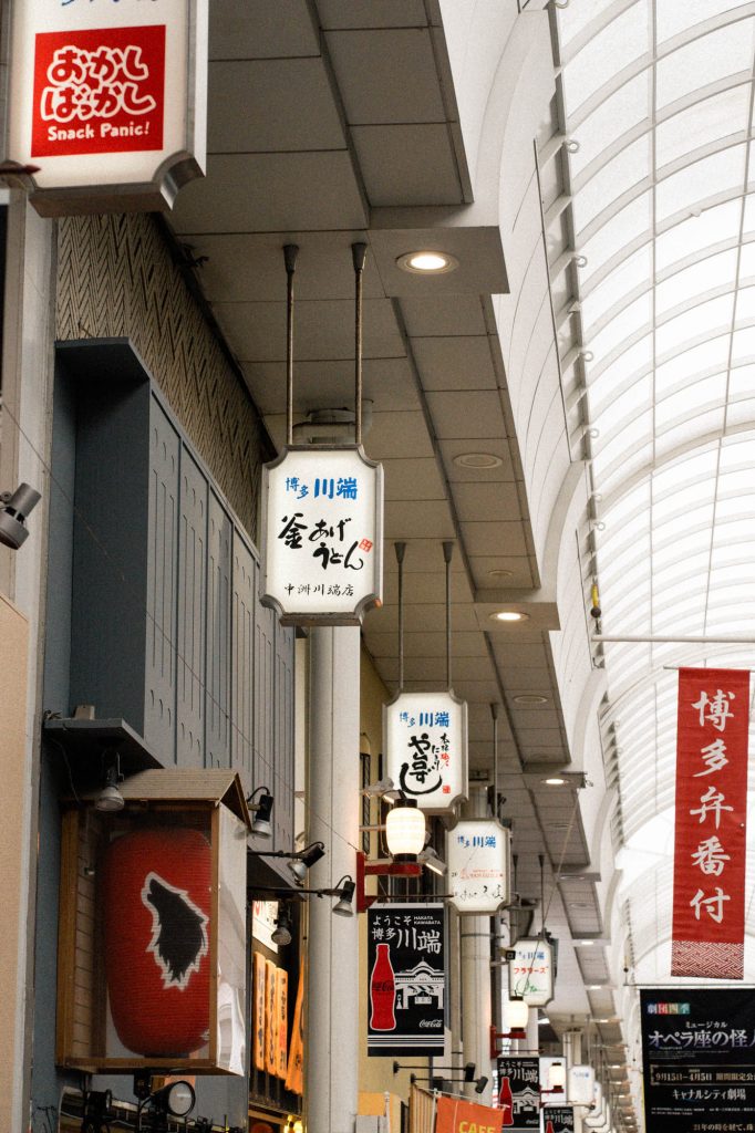 上川端商店街のアーケード内に並ぶ、「博多川端」「釜あげうどん」などの白い吊り看板と、赤い「博多弁番付」の旗が続く奥行きのある風景。