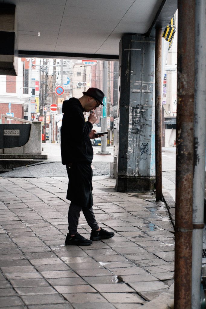 福岡市博多区、雨上がりの路上でハットを被り、スマートフォンを操作しながら電子タバコを吸う男性。背景には「冷泉橋」の文字や街頭が見える、冷たい質感のスナップ。