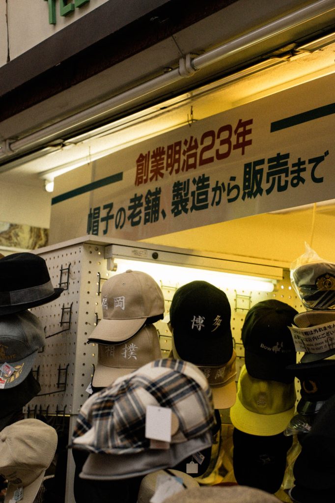 明治23年創業の帽子の老舗店の陳列棚。福岡や博多、相撲といった文字が刺繍されたキャップが並び、上部には「帽子の老舗、製造から販売まで」という看板が見える。