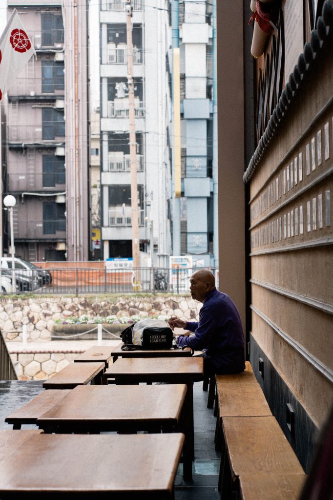 博多川沿いのテラス席に座る年配の男性。背景には中洲のビル群がそびえ立ち、静かな都会の日常を切り取った風景。