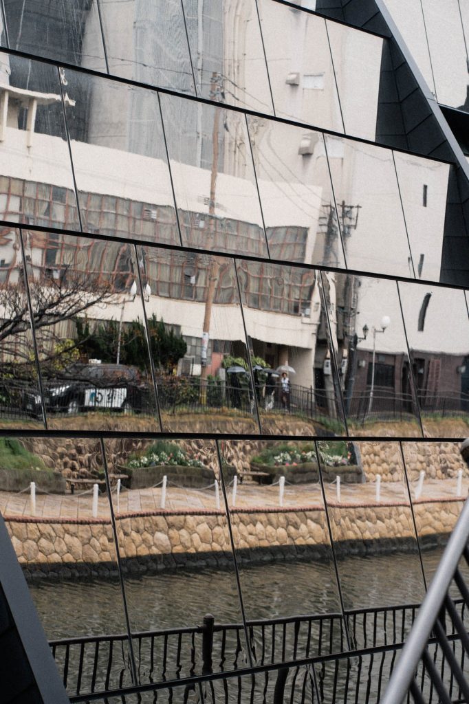 ガラス張りの建物に反射して映し出された博多川と対岸の風景。傘を差して歩く人々のシルエットが歪んで写り、抽象的な視覚効果を生んでいる。