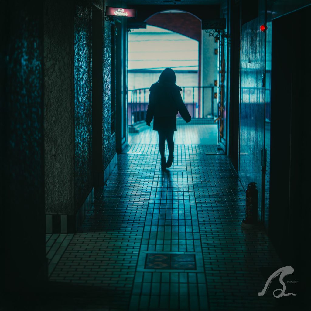 福岡市博多区中洲にあるレトロビル「丸源36ビル」の内観廊下で、モデルの遥香さんの後ろ姿を捉えたシルエットポートレート。 青みがかった暗い廊下の両側に質感のある壁が続き、床には細かなタイルが敷き詰められています。廊下の先にはアーチ状の出口があり、そこから眩い光が差し込んでいます。 画面左上には「BAR Velour」の小さな看板、右下には消火器が配置され、中洲のビルの日常と非日常が混ざり合う雰囲気を表現しています。右下にはPhotossierのロゴが入っています。