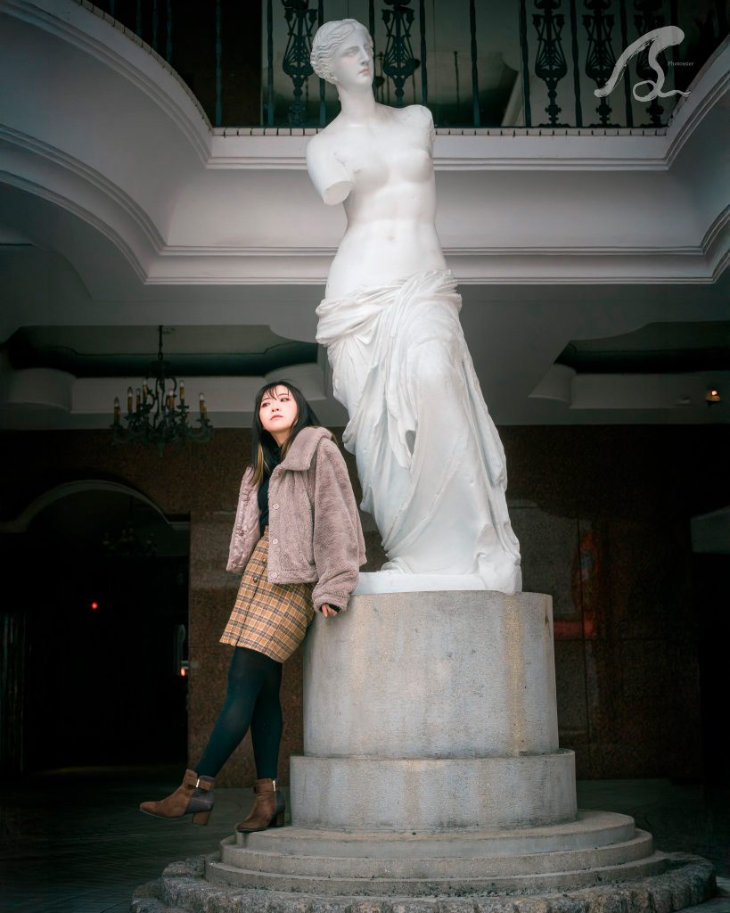 福岡市博多区中洲の丸源36ビルロビーにて、巨大なミロのヴィーナス像の台座に寄り添うモデルの遥香さんのポートレート。 遥香さんは、茶色のボアジャケットに黄色のチェック柄ミニスカート、黒タイツ、茶色のアンクルブーツを着用し、斜め上を静かに見つめています。 背景にはクラシックなデザインのシャンデリアや、曲線が特徴的な白いバルコニーが見え、重厚でアンティークな雰囲気が漂っています。全体的に落ち着いたライティングで、右上にPhotossierのロゴが入っています。