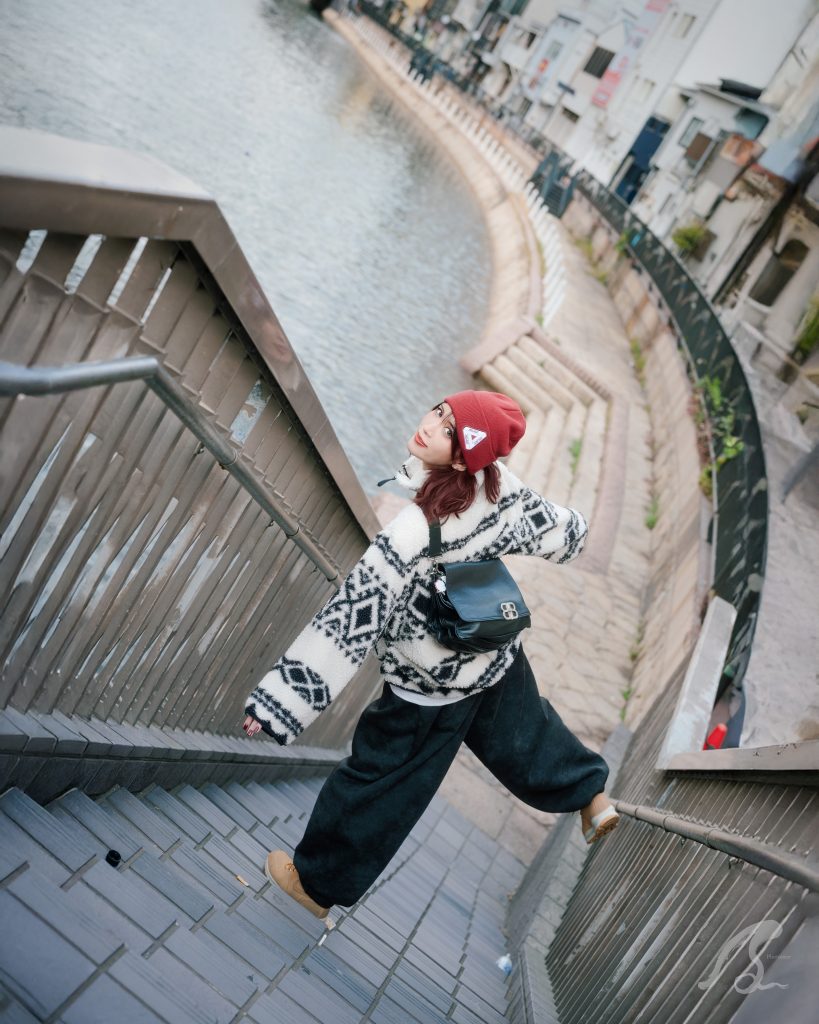 福岡市博多区、川端商店街横の博多川沿いにある階段で、モデルのHonopiさんを撮影したストリートポートレート。 Honopiさんは、白と黒の幾何学模様が入ったボアジャケットに、黒のゆったりとしたワイドパンツ、ベージュのスニーカー、そして赤いニット帽を着用しています。彼女は階段を下りながら、カメラの方を振り返り、印象的な表情を見せています。 背景には穏やかに流れる博多川と対岸の建物が並び、都市の川辺らしい風景が広がっています。画面右下にはPhotossierのロゴが入っています。