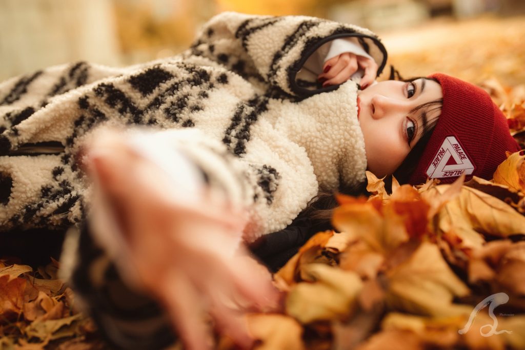 福岡市博多区の冷泉公園にて、地面に敷き詰められた茶色やオレンジの落ち葉の上に横たわるモデルのHonopiさんのクローズアップポートレート。 Honopiさんは白と黒の幾何学模様のボアジャケットに、赤い「PALACE」のロゴ入りニット帽を着用しています。彼女の瞳にピントが合い、手前にある手や落ち葉は大きくボケており、写真に深い奥行きを与えています。 背景には秋の午後の柔らかな光が差し込み、全体的に暖色系でまとめられた、しっとりとした秋の空気感を感じさせる1枚です。右下にはPhotossierのロゴが入っています。