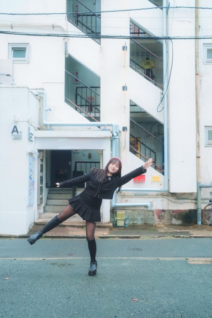 福岡市博多区にある登録有形文化財「冷泉荘」の白い建物を背景に、モデルのぴよさんがポーズをとる全身ショット。 ぴよさんは黒のツイード風セットアップ（ジャケットとミニスカート）に黒のタイツと厚底ブーツを着用し、赤いカチューシャをしています。片足を上げ、両腕を翼のように広げた躍動感のあるポーズで笑顔を浮かべています。 背景は、昭和レトロな雰囲気の集合住宅で、特徴的な外階段や淡いブルーの露出配管、左側には「A棟」と記された看板が見えます。全体的に明るく柔らかな自然光に包まれた写真です。