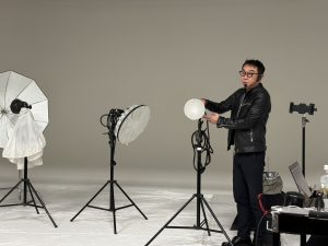 薄暗いスタジオで、革ジャン姿の笹口氏が、様々な照明機材（アンブレラ、ソフトボックスなど）を調整している。