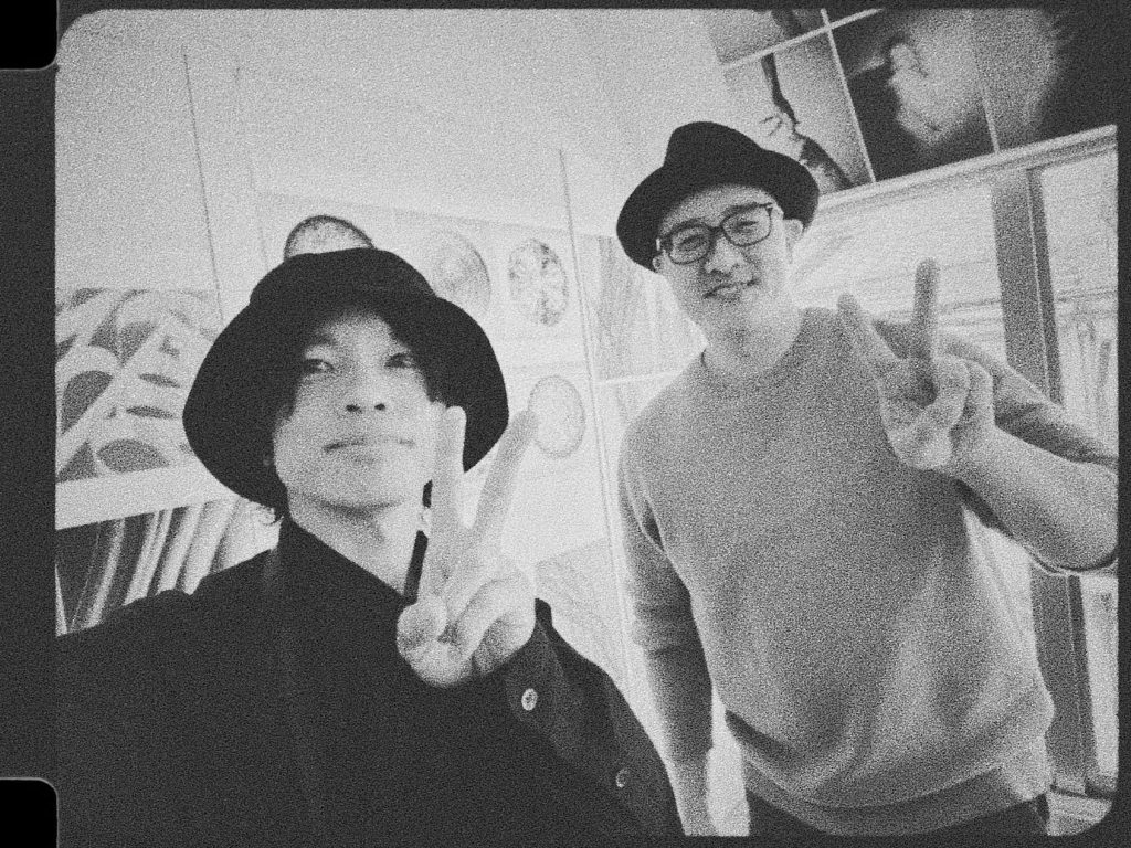 バケットハットのイソ氏と中折れ帽の赤木友厚が並び、笑顔でピースサインをするモノクロ写真。