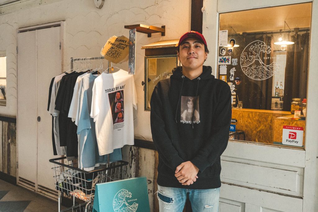 入口付近のTシャツラックの横で、穏やかな表情で立つ西本さん。