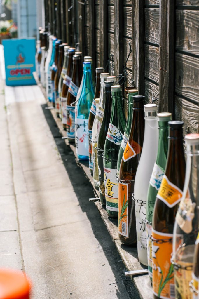 大名の路地裏に整然と並べられた焼酎や日本酒の一升瓶。日差しが反射し、背景がボケている。