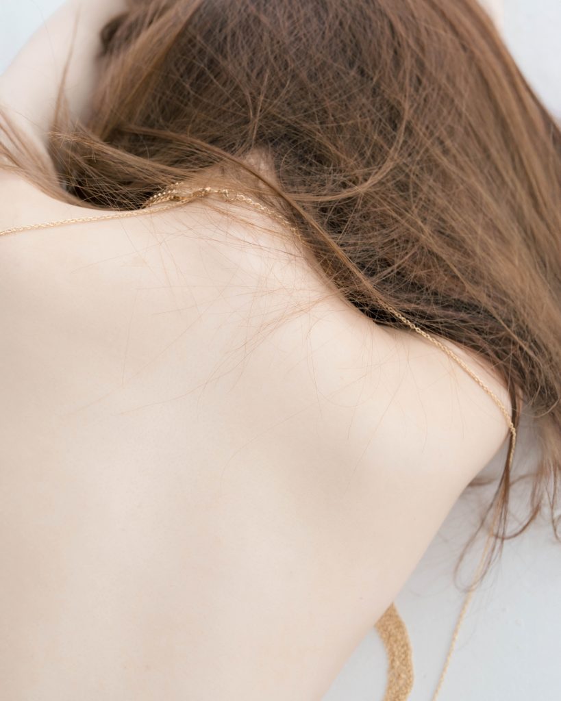 女性の背中からうなじにかけての接写。柔らかな髪の毛と肌の質感が、明るい光の中で繊細に描写されている。