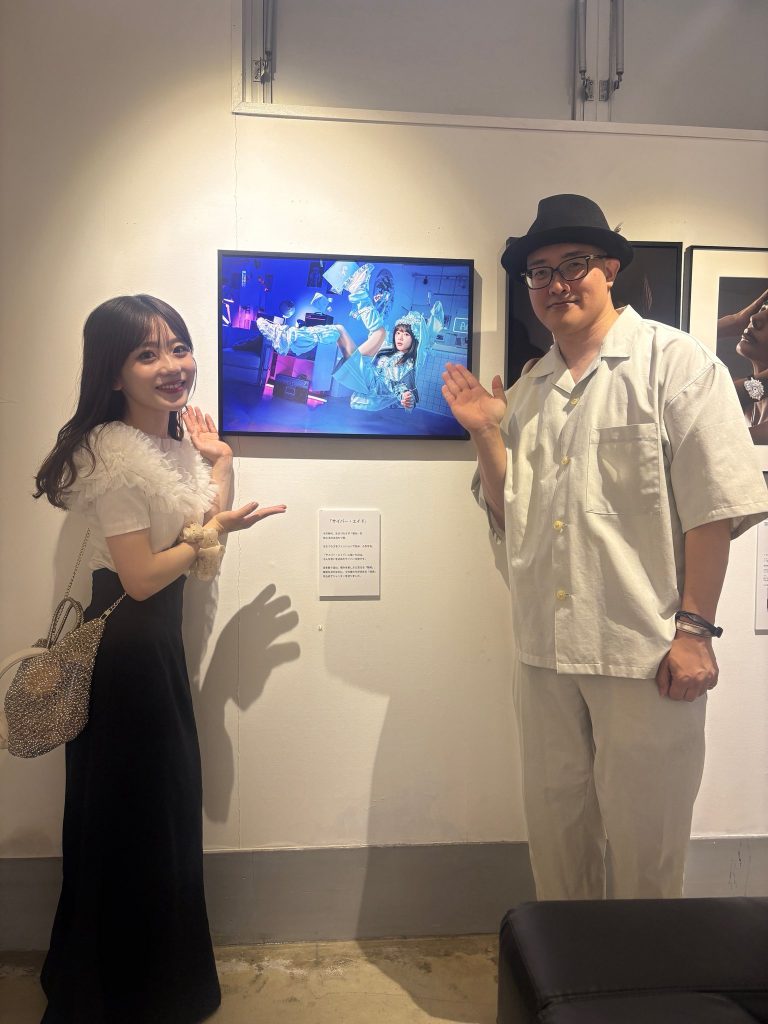 福岡のアートギャラリーART BASE KMYの写真展会場で、赤木友厚の写真作品「サイバー・エイド」の隣に、モデルを務めたこもれびもかさんと赤木友厚が並んで立っている。こもれびもかさんは作品と同じ、両手でハートマークを作るポーズをして笑顔。赤木友厚も隣で笑顔。