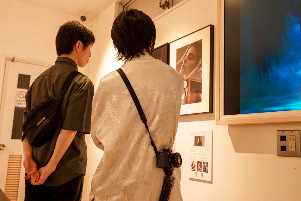 ギャラリーの壁に展示されたポートレート作品を真剣な表情で見つめる、カメラを背負った2人の若い男性の後ろ姿