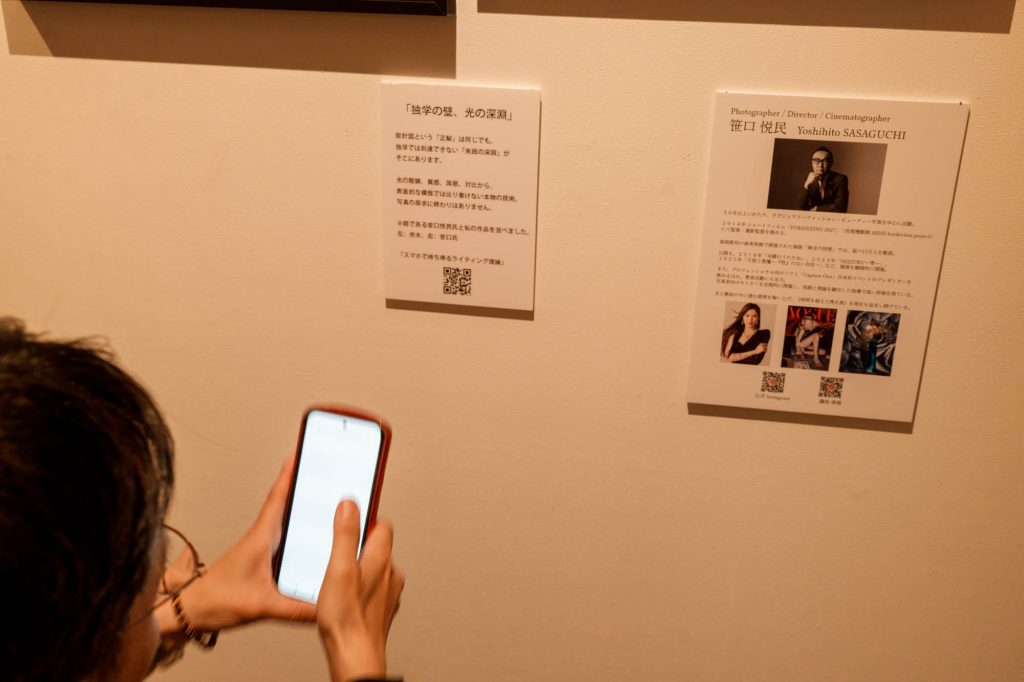 壁に貼られた赤木友厚の「独学の壁、光の深淵」キャプションのQRコードを、来場者がスマートフォンで読み取っている手元のアップと、隣にある写真家・笹口悦民のプロフィールパネル
