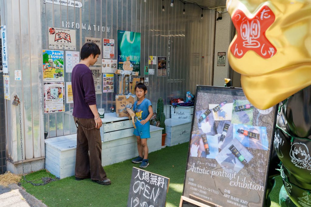 ART BASE KMYの人工芝エリアで、写真展の案内看板の奥に立ち、ダンボールの工作を前に話をしている男性と青い服を着た地元の男の子