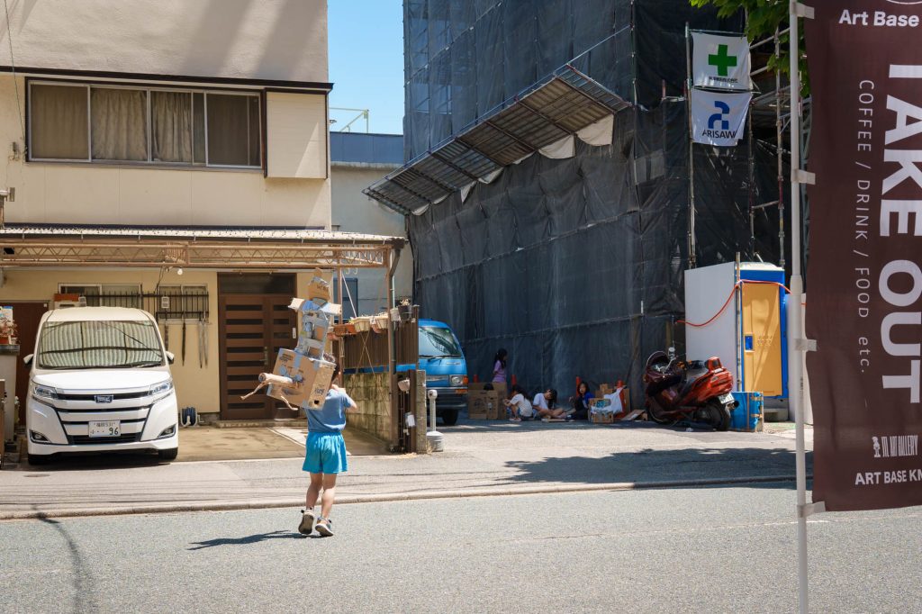 頭からダンボールで作った大きなロボットのような工作を被り、青いTシャツとハーフパンツ姿で道路を歩いていく男の子の後ろ姿