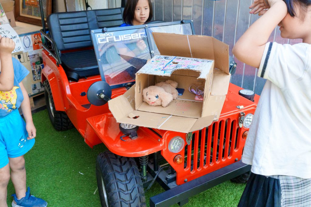 人工芝の上に停められた赤いミニカー(next CRUISER)と、そのボンネットに置かれたダンボール箱の中に大切そうに収められた犬のぬいぐるみ